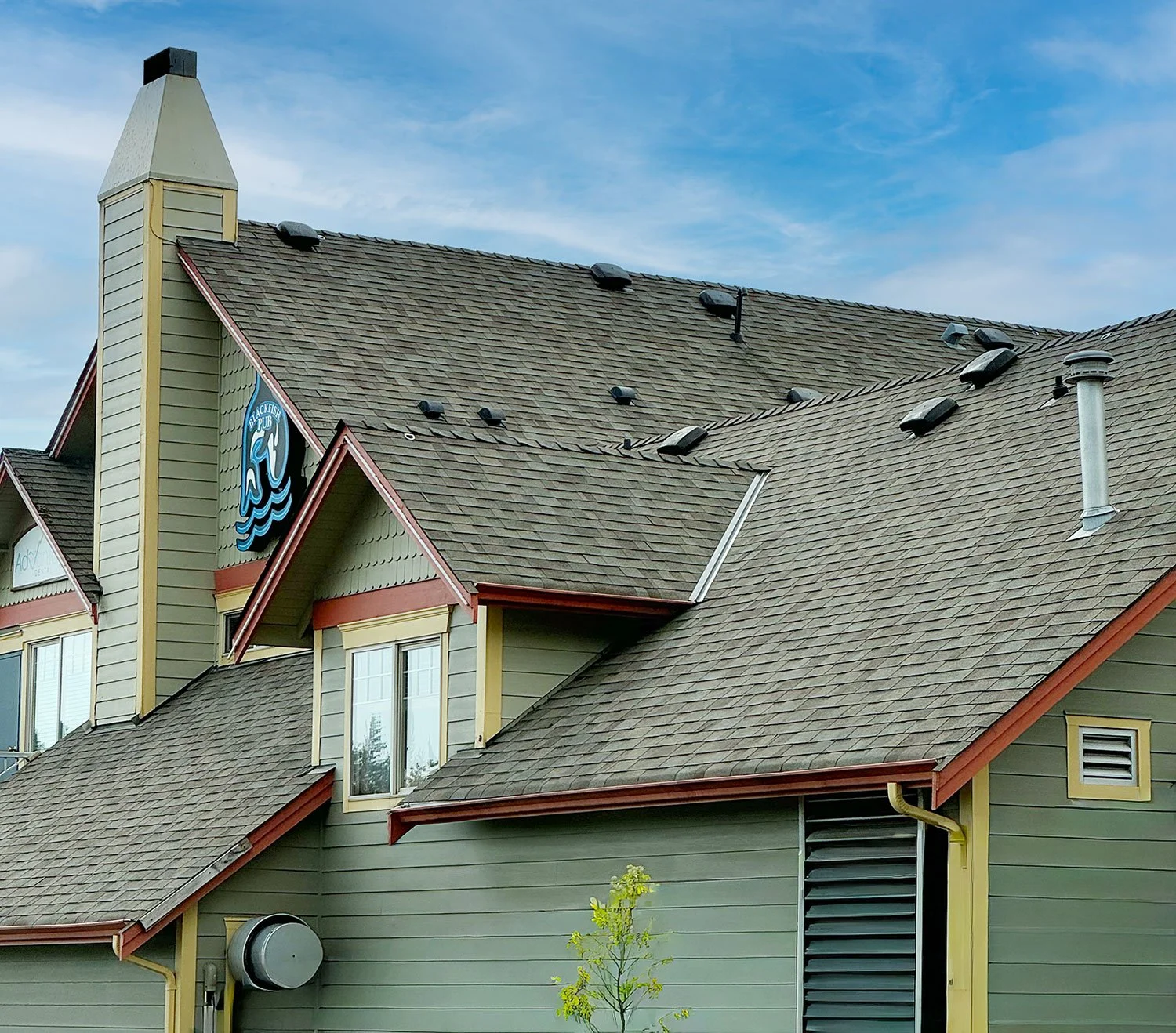 A residential building with green siding, multiple roof angles, and black roof vents, under a partly cloudy sky.