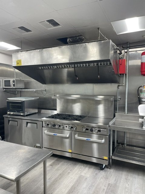 Commercial kitchen with stainless steel stove, oven, range, exhaust hood, microwave, and shelving.