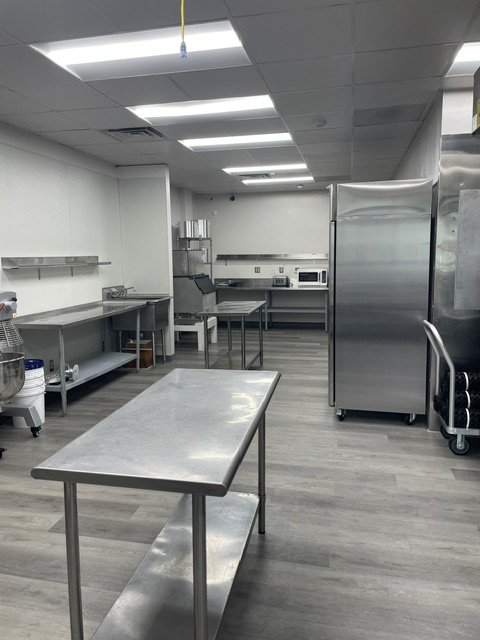 Empty commercial kitchen with stainless steel tables and appliances, including a large refrigerator, microwave, and storage shelves.