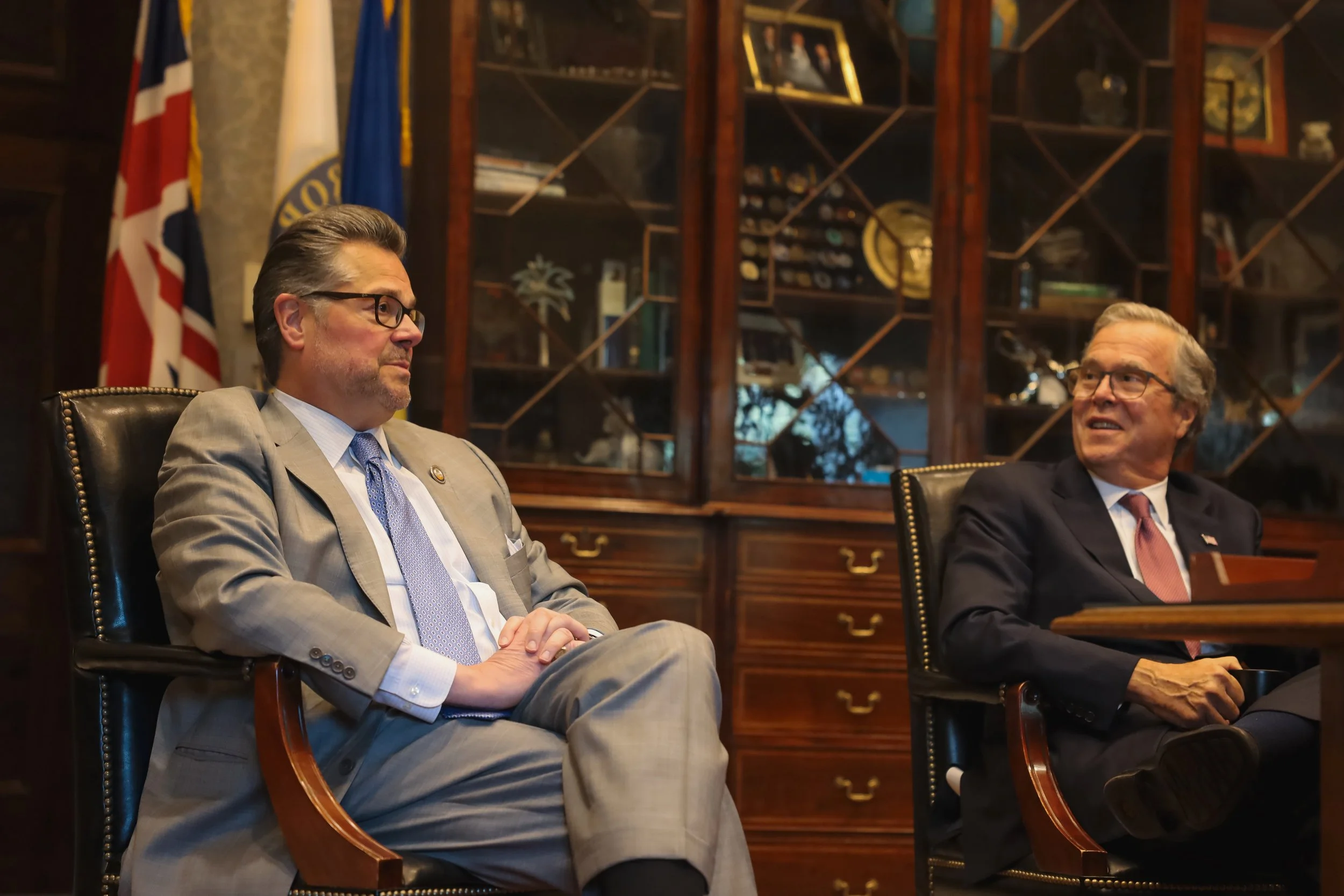 Two men in suits sitting at a conference table in a formal setting, with a bookcase and flags in the background.