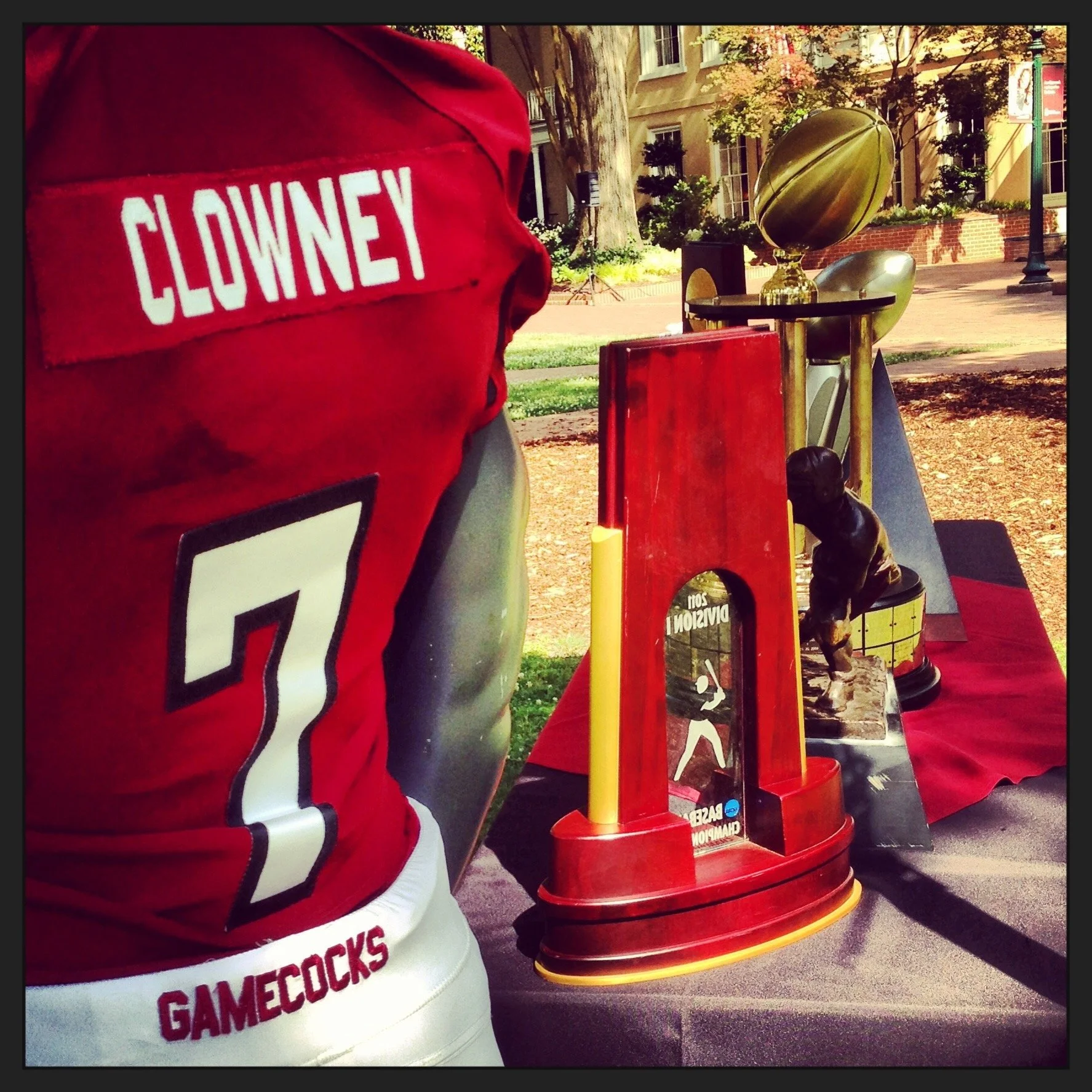A street scene with a focus on a person wearing a red jersey with 'CLOWNEY' written on the back and the number 7, standing near a display of the Vince Lombardi Trophy, which is placed on a table with a red tablecloth and surrounded by various signs and items, with trees, buildings, and a lamppost in the background.