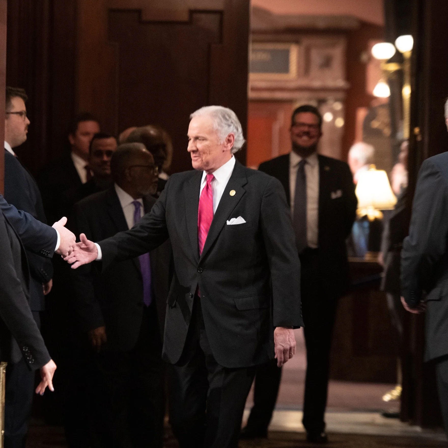 A man in a black suit with a pink tie shaking hands with another man in a dark suit at an indoor event, with several other men in suits standing nearby, some smiling, in a dimly lit room with warm lighting and wooden paneling.