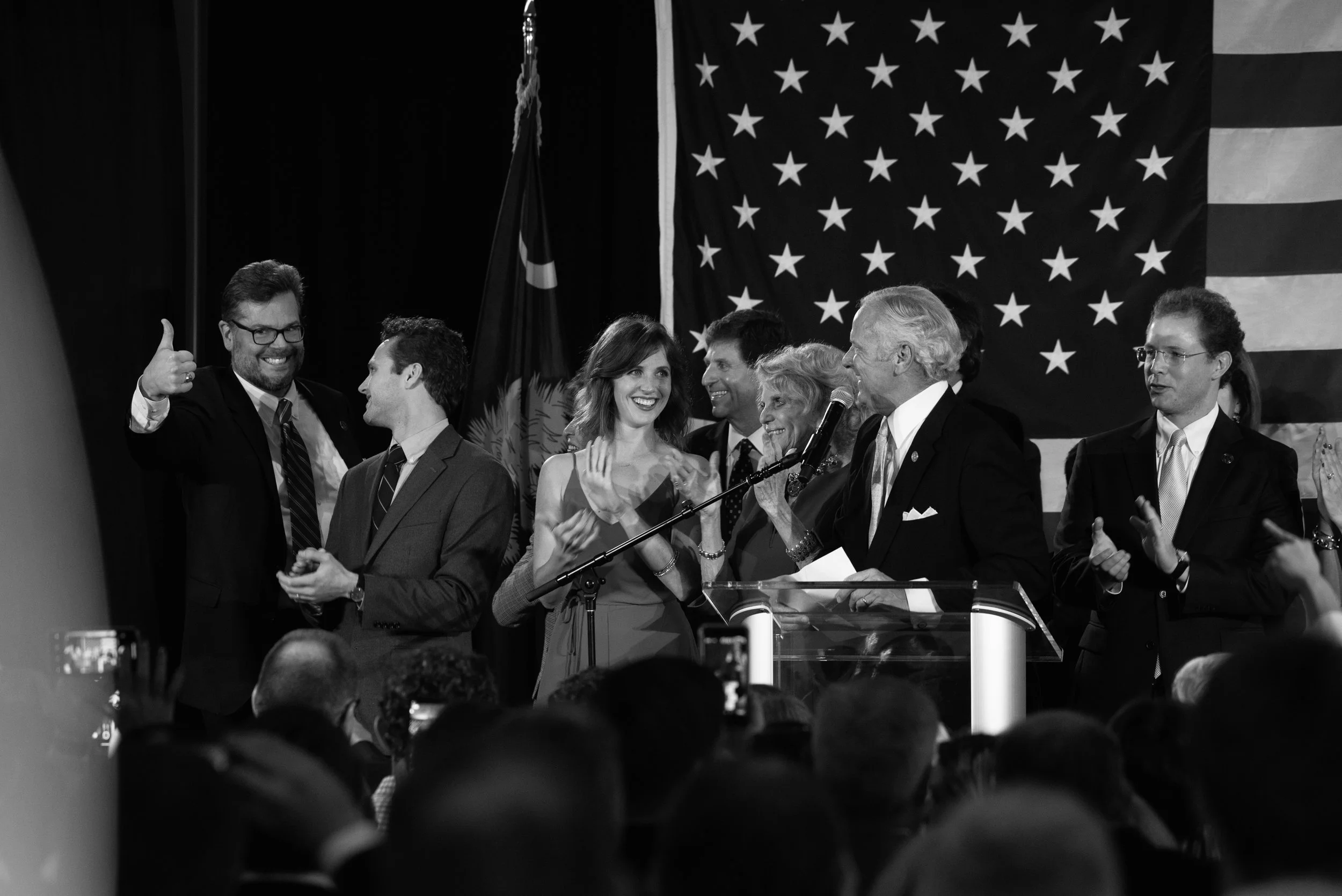 Black and white photo of a group of people on stage at a political event, with an American flag and South Carolina state flag in the background.