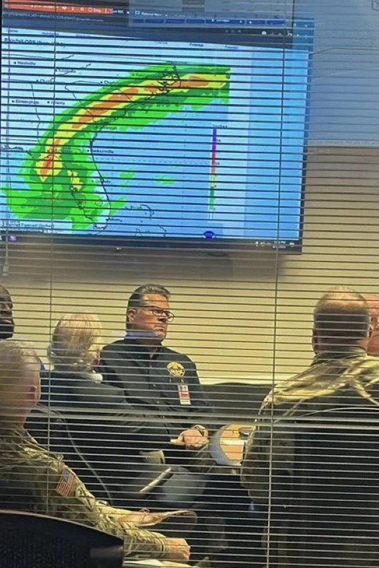 A weather forecast meeting shows a large weather map with a storm system, likely a hurricane, depicted with red, yellow, and green colors. The team, including a man in a black uniform and others in military attire, is seated behind horizontal blinds observed from outside the room.