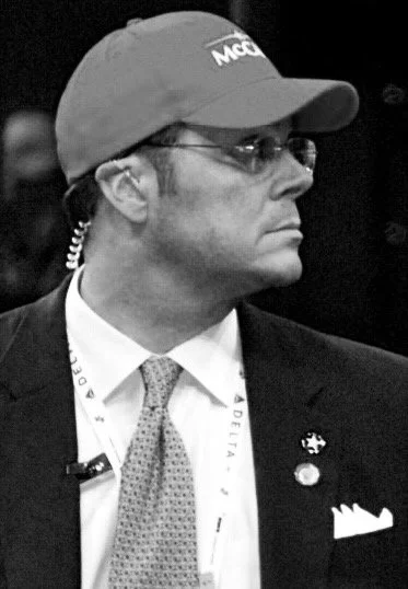 A man wearing glasses, a baseball cap with 'McDonald's' logo, a suit jacket, dress shirt, and tie, with various pins and a lanyard.