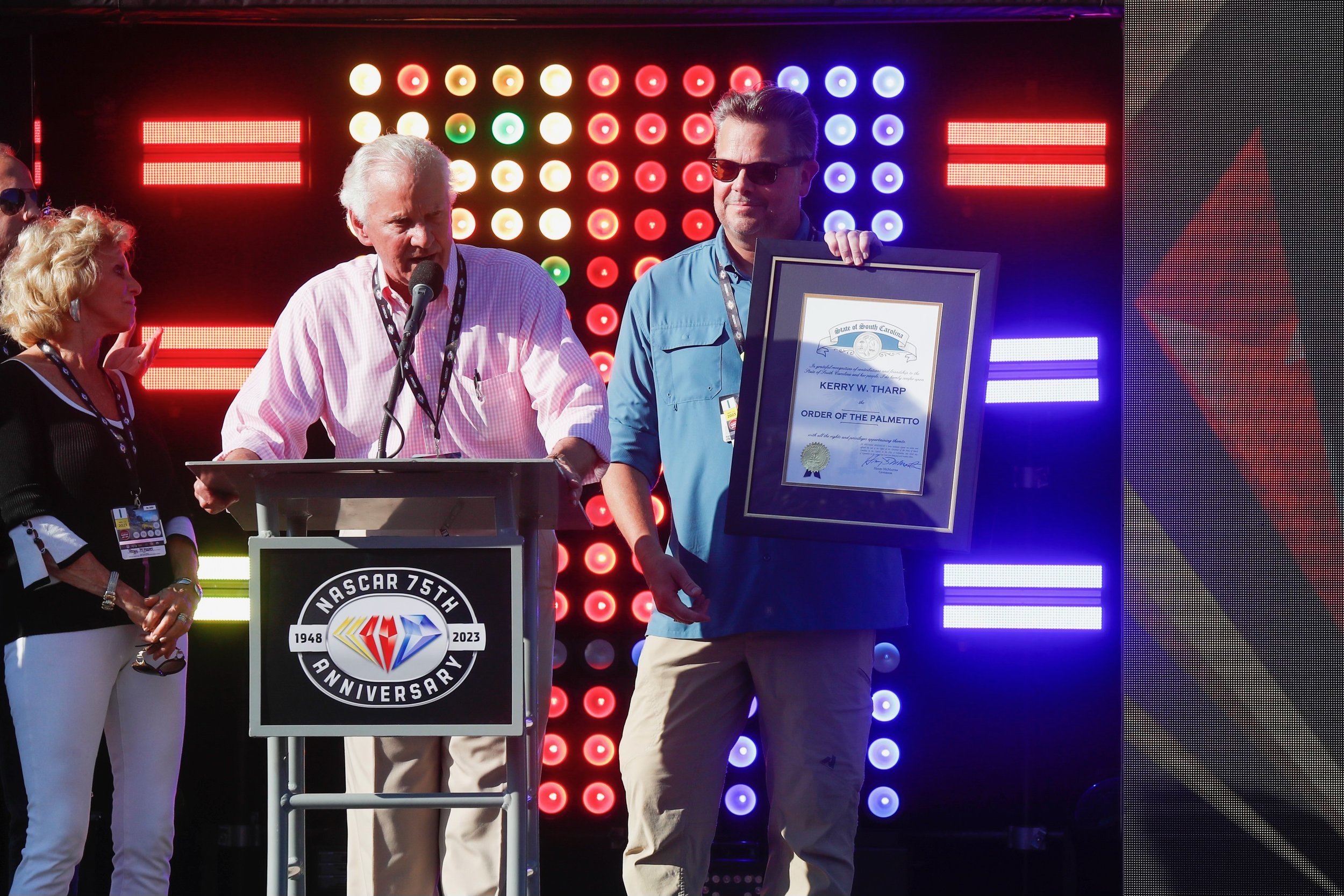 A man in a pink shirt speaking at a podium with a NASCAR 75th anniversary logo, an older man with white hair holding a framed certificate, and a woman with blonde hair watching, at a NASCAR event with colorful LED lights in the background.