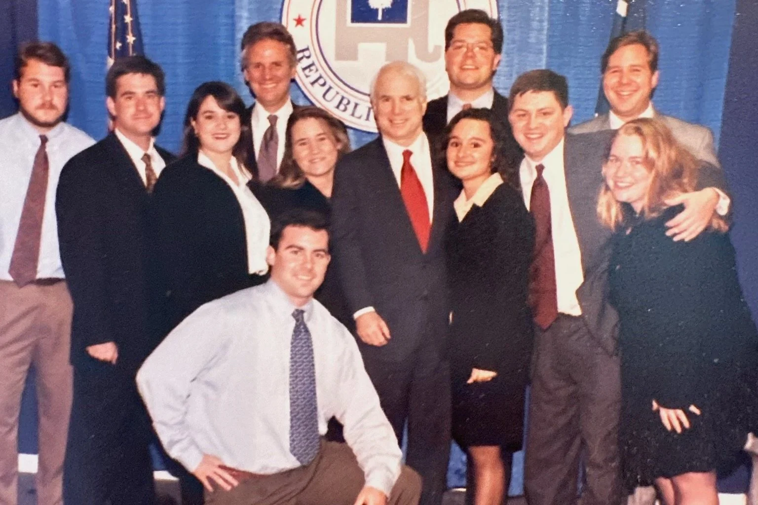 A group of 12 people, mostly young adults, dressed in business attire with suits and ties, posing together at a formal event in front of a blue curtain with a logo and the text 'REPUBLIC' partially visible.