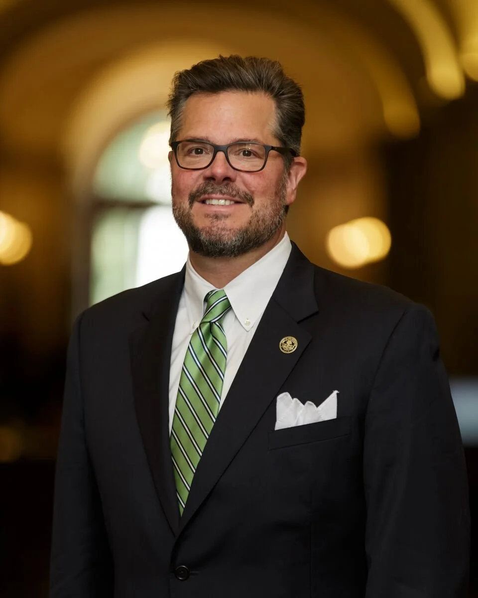 A middle-aged man with glasses and a beard, wearing a black suit with a white shirt and a green striped tie, standing indoors with a blurred background.