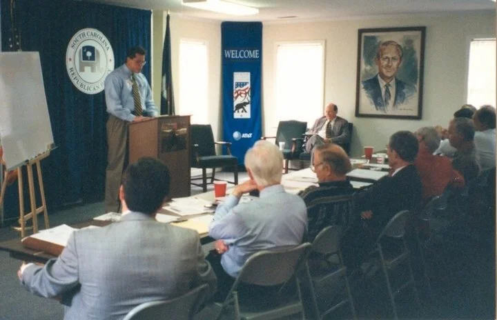 A man speaking at a podium in a room with a group of people seated at tables, some taking notes, with South Carolina state symbols and a portrait hanging on the wall.