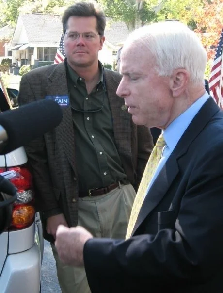 Two men, one older with white hair in a dark suit and yellow tie, the other younger with dark hair and glasses in a green shirt and blazer, stand outdoors near a car during a discussion or interview.