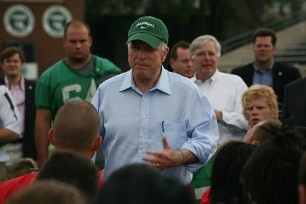 A man wearing a green baseball cap and a light blue button-up shirt is speaking to a group of children with adults standing behind him.