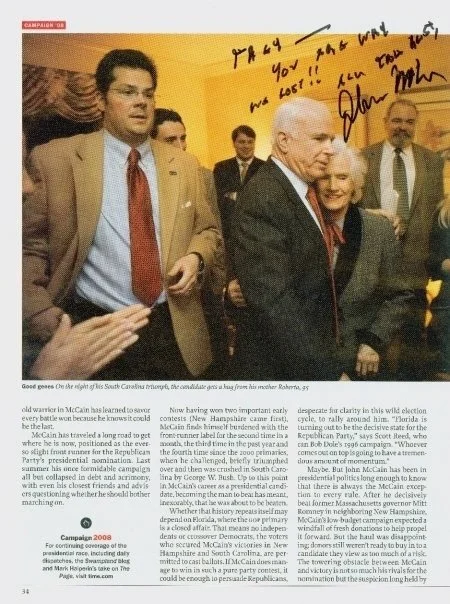 Group of men at a political event, with one man signing an autograph on a yellow note, surrounded by others in suits, in what appears to be a campaign gathering.