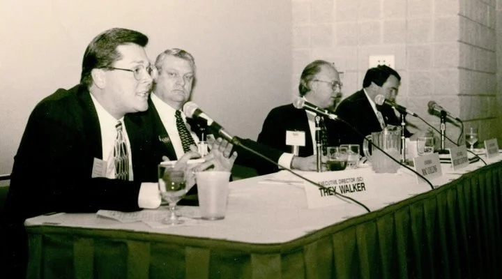 Five people sitting at a panel discussion table with microphones, glasses of water, and name tags, in a conference room.