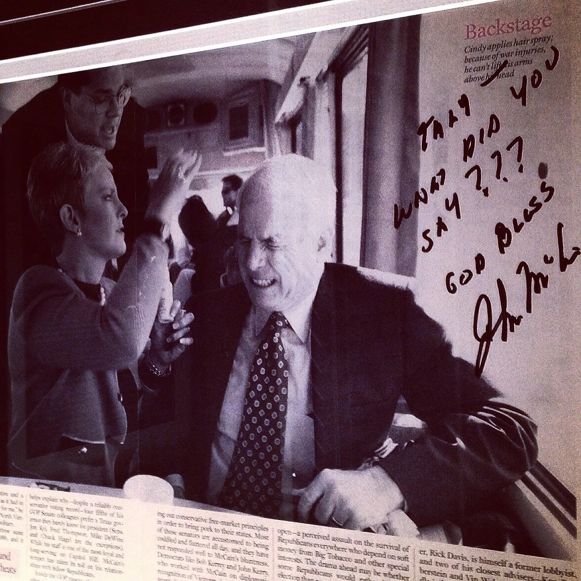 Black-and-white photograph of a woman and an older man smiling and talking at a table, with a man standing behind them. Handwritten notes are on the photo, asking about how to say something and mentioning a press. The top right corner has a typed caption that references backstage hair spray and arm injuries.