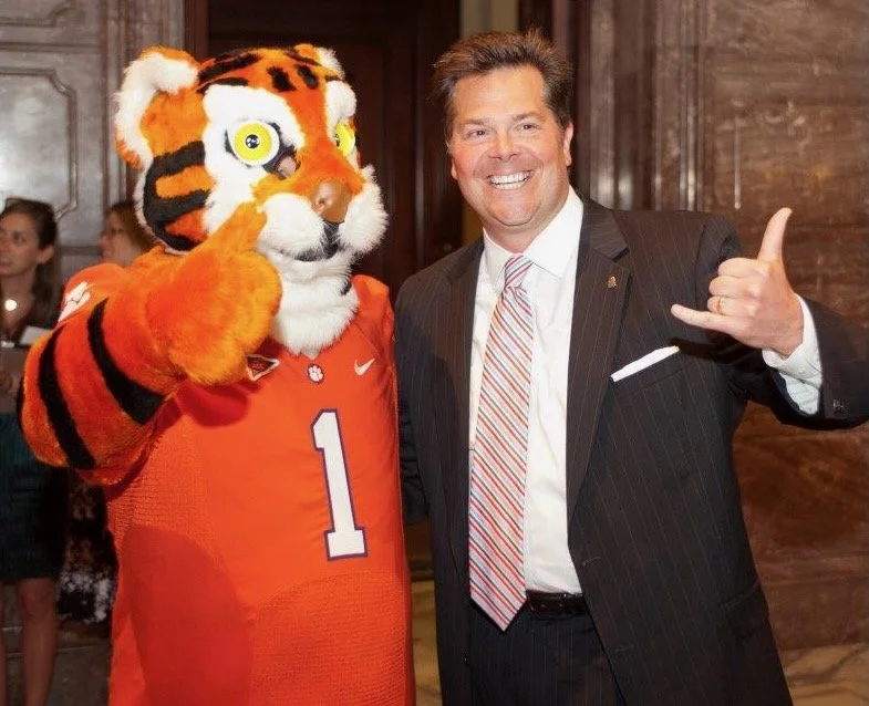 A man in a black suit and striped tie smiling and giving a shaka sign next to a person in a tiger mascot costume wearing an orange sports jersey with the number 1, in a room with wooden walls, some people in the background.