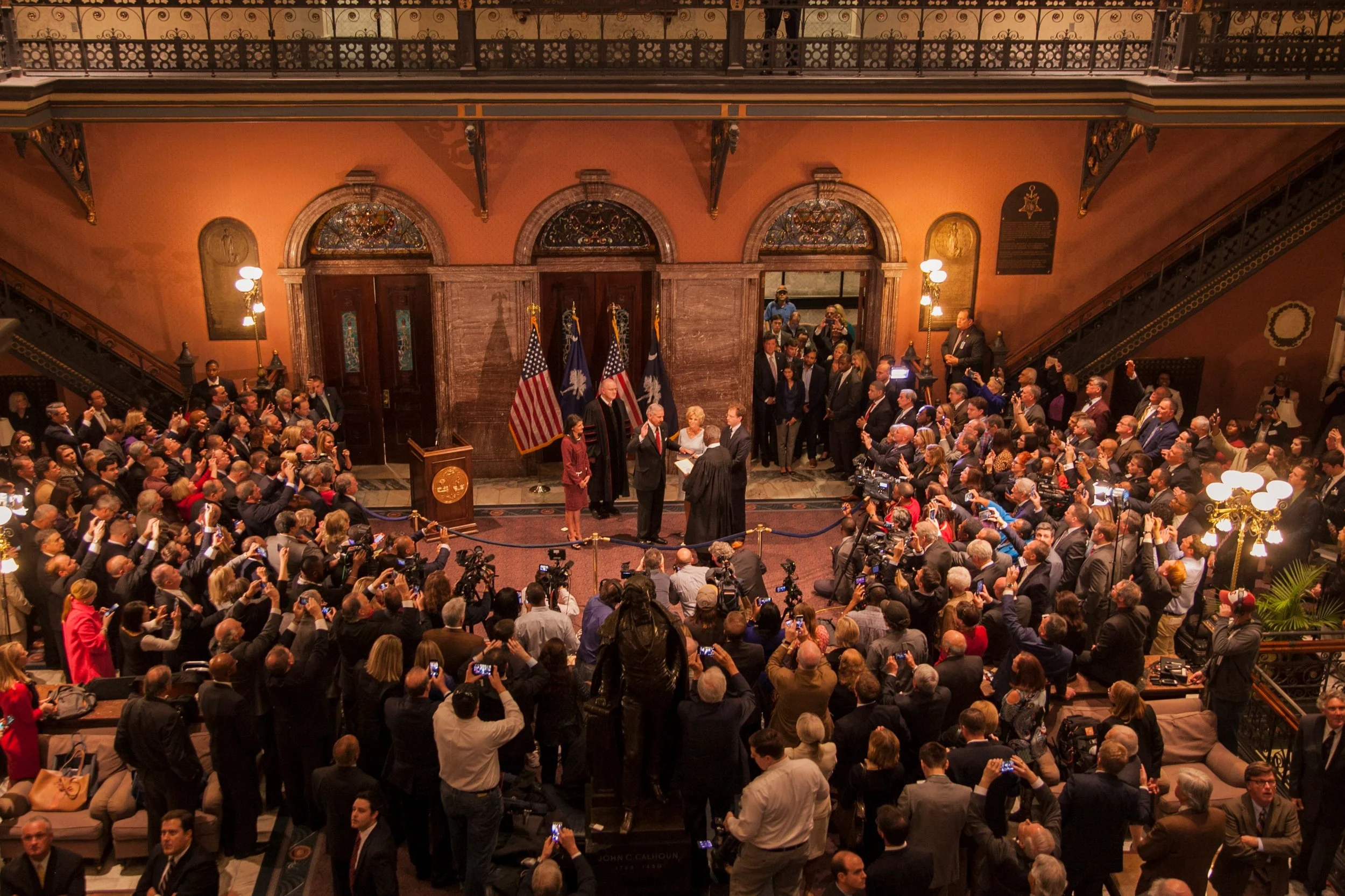 A formal government swearing-in ceremony with officials and a crowd seated in an ornate room decorated with flags and artwork.