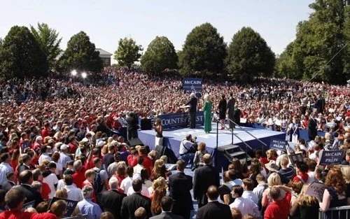 Large outdoor rally with a stage, speaker, and a crowd of supporters, trees in the background, and signs supporting McCain and Palin.
