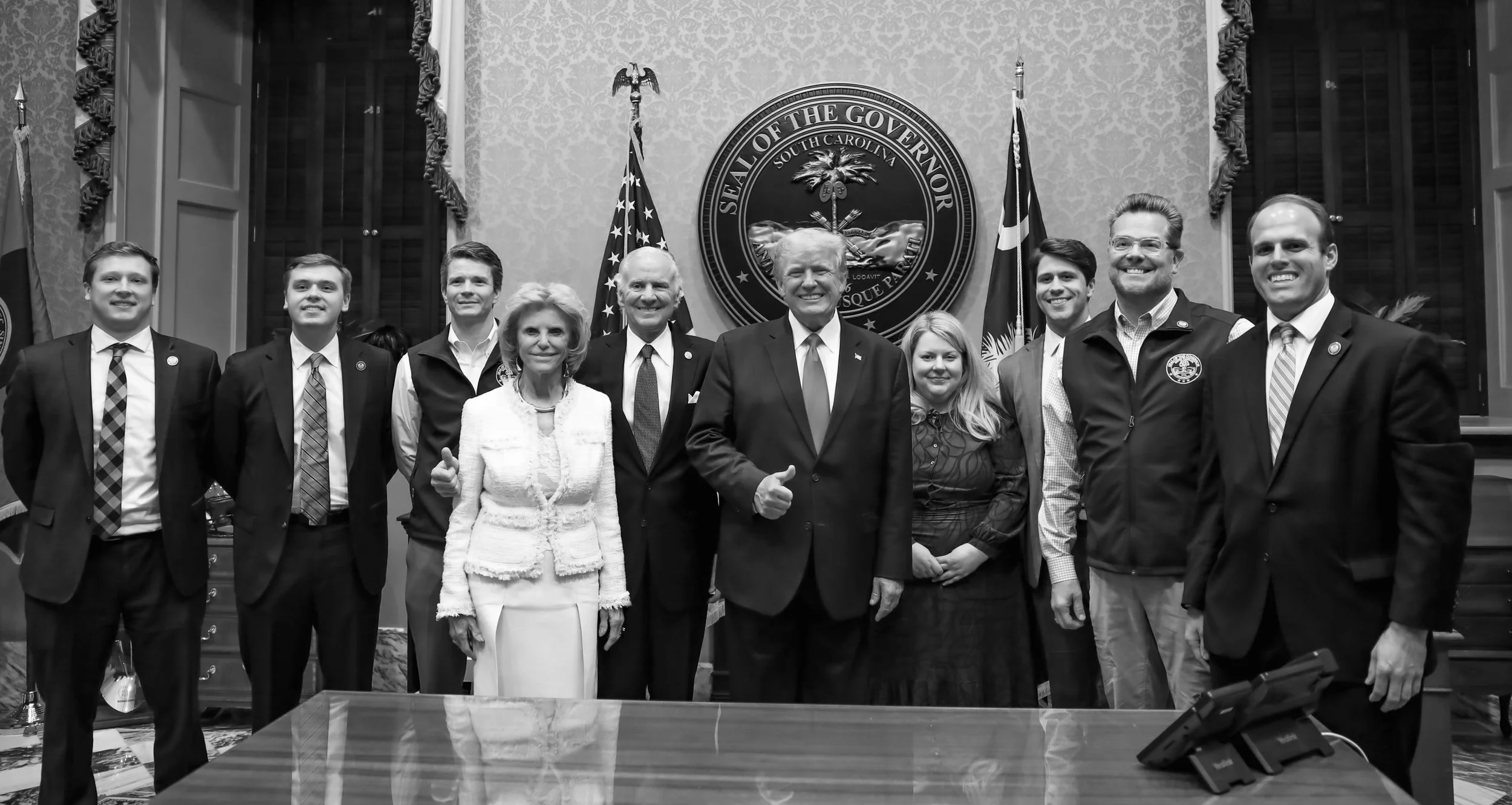 A group of nine people standing together in a formal setting, with flags and a seal that reads 'Seal of the Governor South Carolina' in the background.