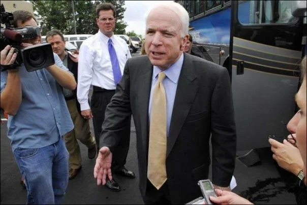 A man in a black suit and yellow tie speaking to reporters outside, with several people, some holding cameras and microphones, around him.