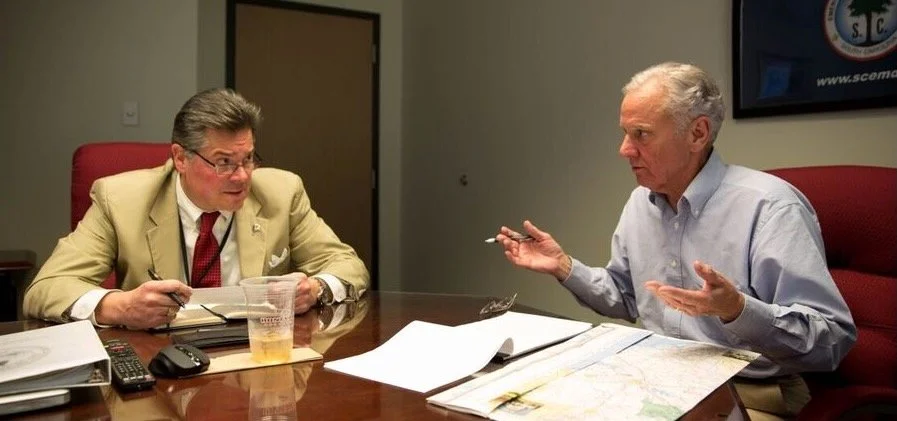 Two men are sitting at a conference table having a discussion. The man on the left wears glasses and a beige suit, holding a pen over a notepad with a drink in front of him. The man on the right has gray hair, wears a light blue shirt, and gestures with his hands. There are papers, a map, a remote, and a TV in the background.