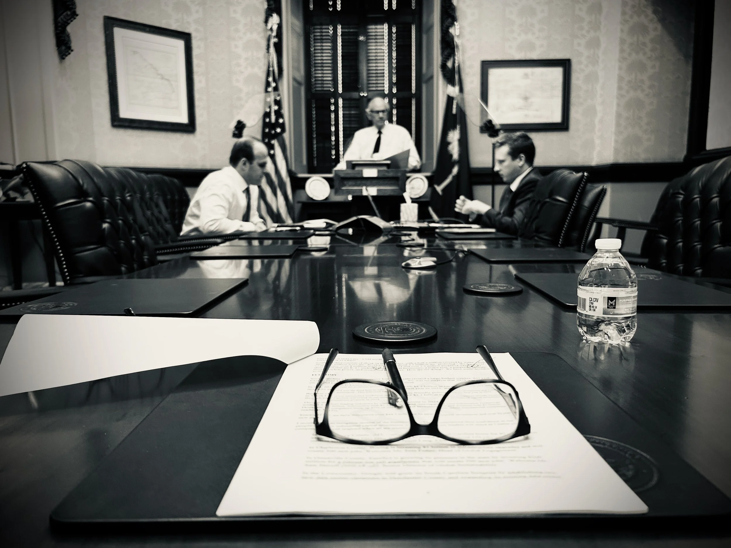 A conference room with empty black leather chairs around a large wooden table. In the foreground, there is a pair of eyeglasses, a pen, and a document on the table. On the table, there is also a plastic water bottle. In the background, three men are seated, paying attention, with another man standing behind a podium. The room has framed pictures on the wall, and American flags are visible. The atmosphere suggests a formal meeting or hearing.