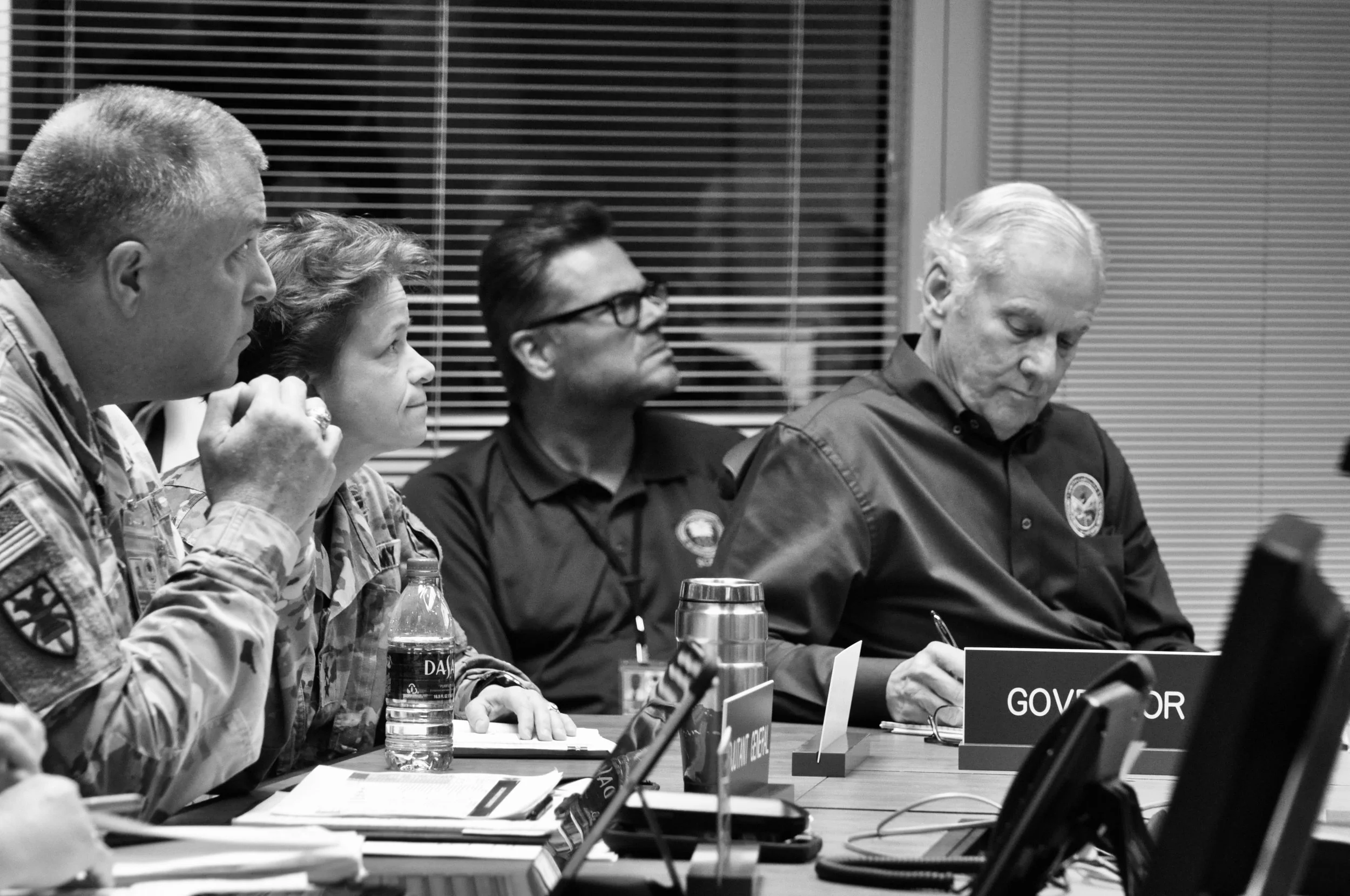 A black and white photo of people sitting at a conference table, with nameplates and documents in front of them. The individuals include a woman, and men wearing uniforms, possibly military or government officials. They are engaged in a serious discussion during a formal meeting.