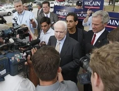 Politician speaking to the media surrounded by reporters and campaign signs that read 'McCain'