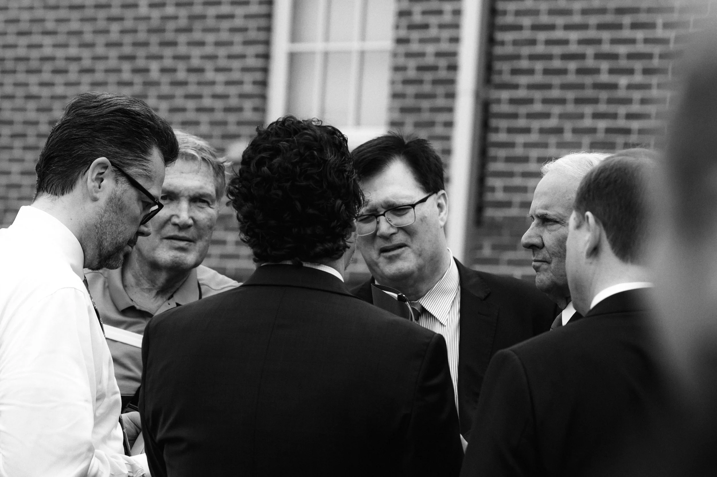 A group of six men standing in a circle engaged in a serious discussion outside, with a brick wall in the background.
