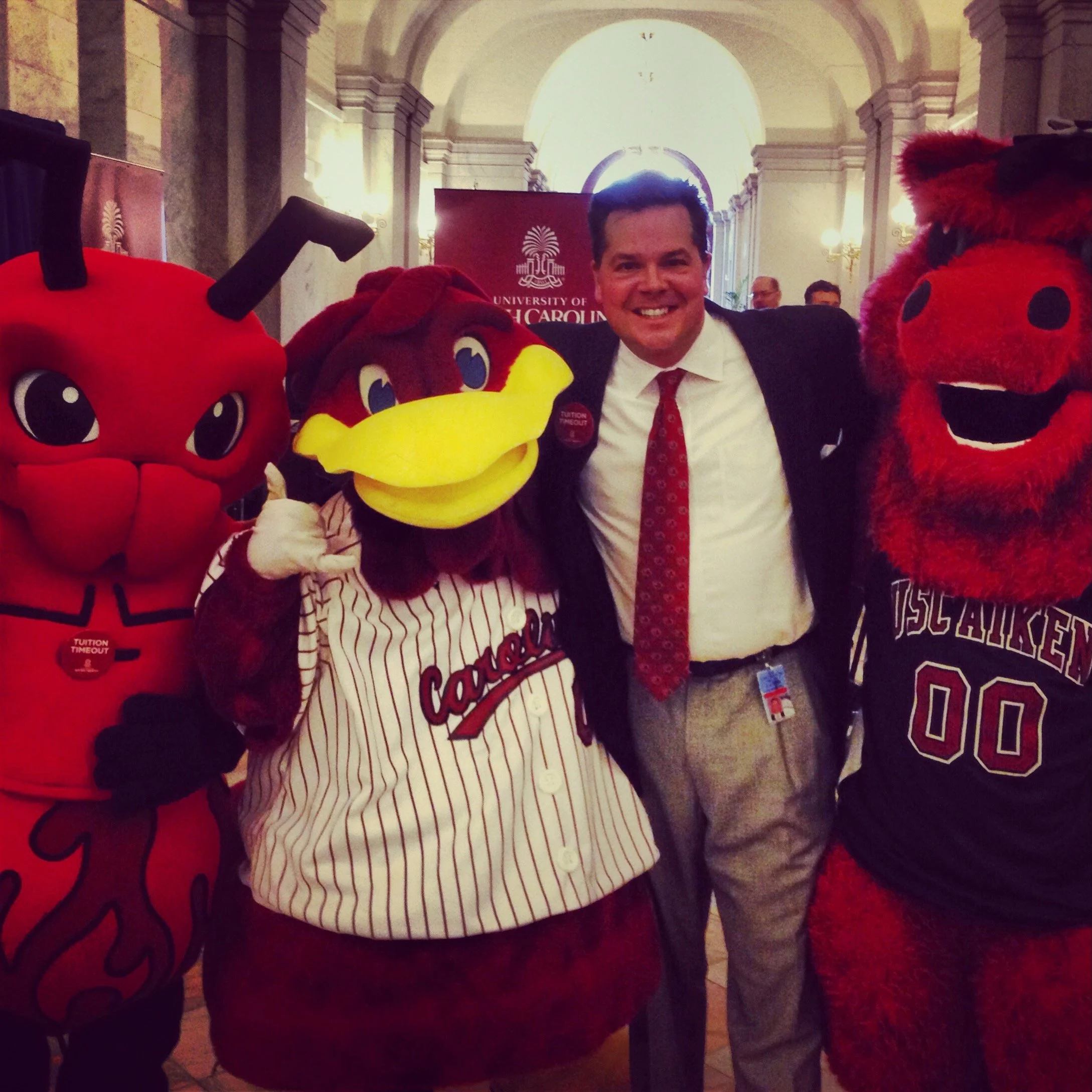 A smiling man in a white shirt, red tie, and black suit, standing with three people in animal costumes in a grand hall with high arched ceilings and ornate lighting. The costumes include a red ladybug, a brown bird with a yellow beak, and a red furry creature with black spots.