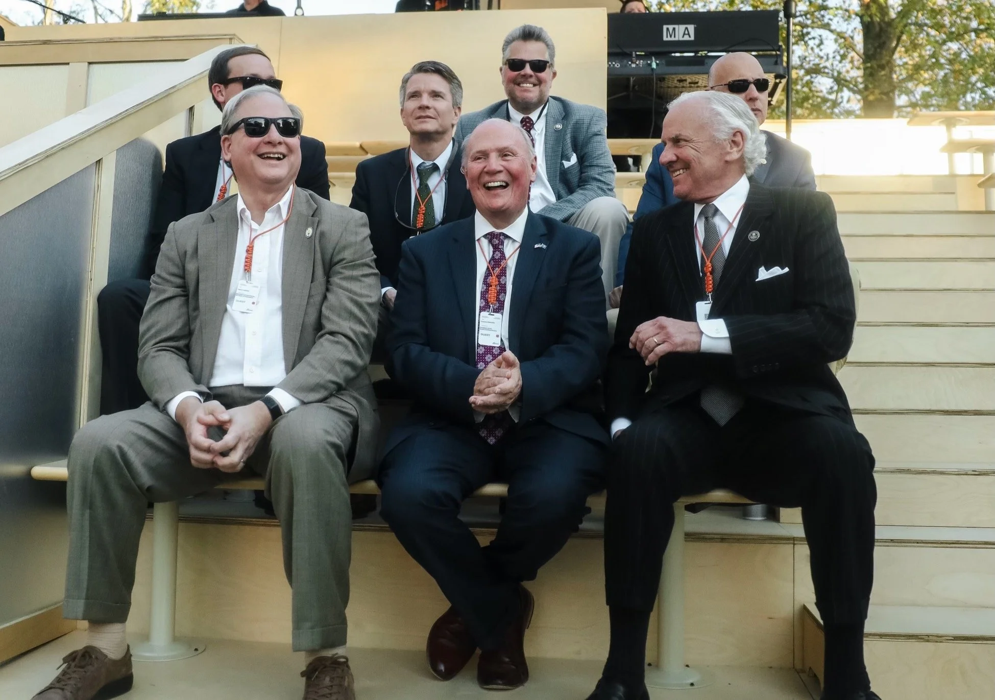 Group of eight men wearing suits, sitting and standing on stairs outdoors, smiling and laughing, some with sunglasses and event badges around their necks.