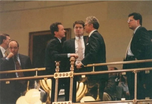 Five men in business attire standing and talking on a gold and black balcony railing in a formal setting.
