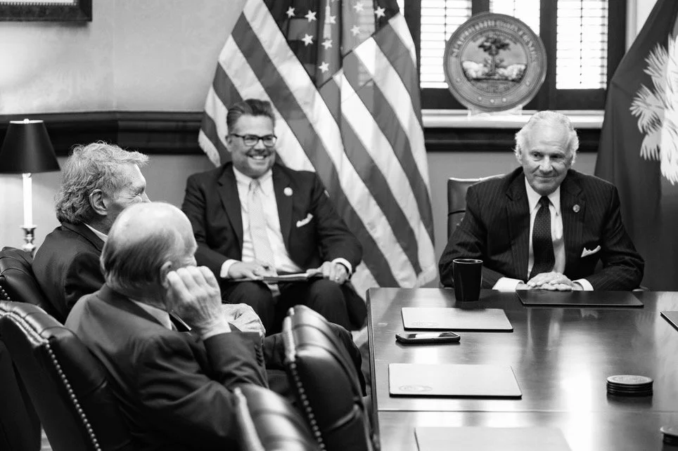 Four men in suits sitting around a conference table with American flags and a South Carolina state flag behind them. One man is smiling, another is speaking, and two are listening. There are laptops and a phone on the table.