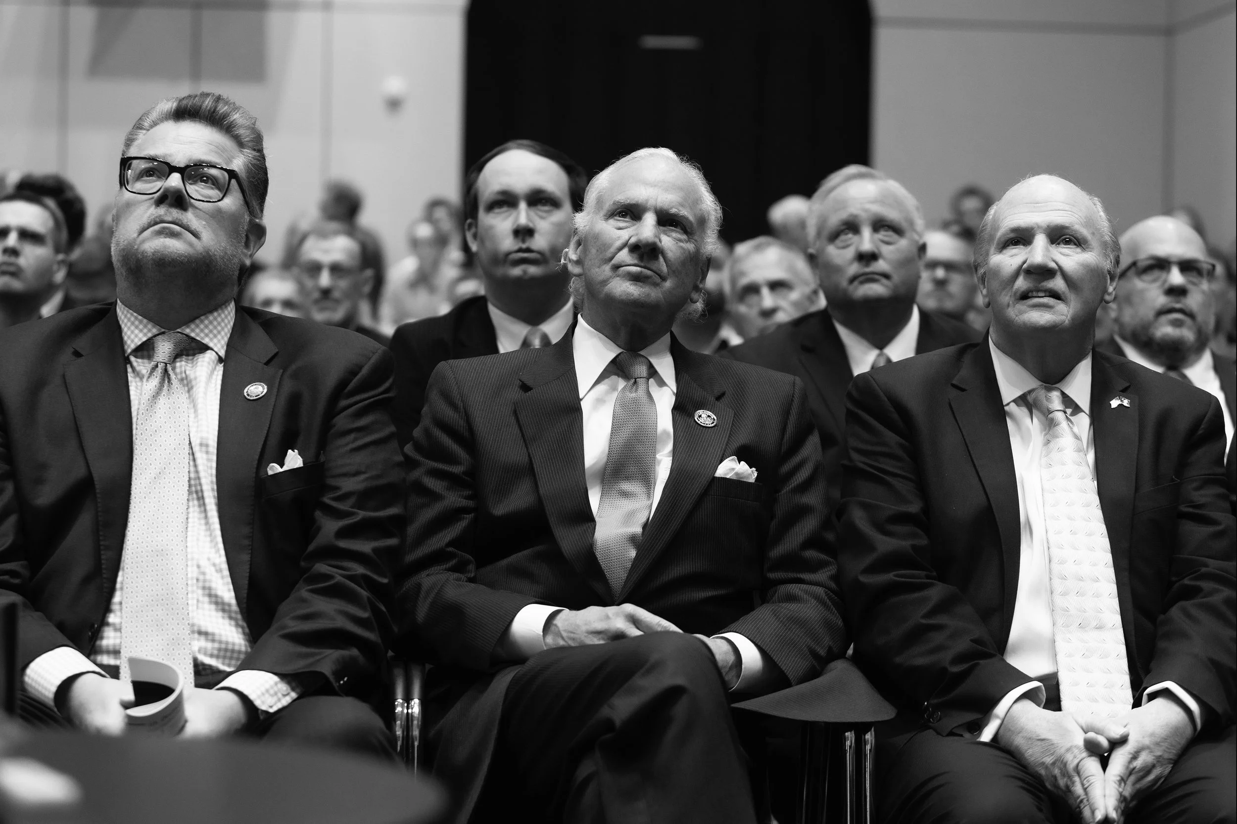 Black and white photograph of a group of men in formal suits attending an event, seated in rows, looking upward attentively.