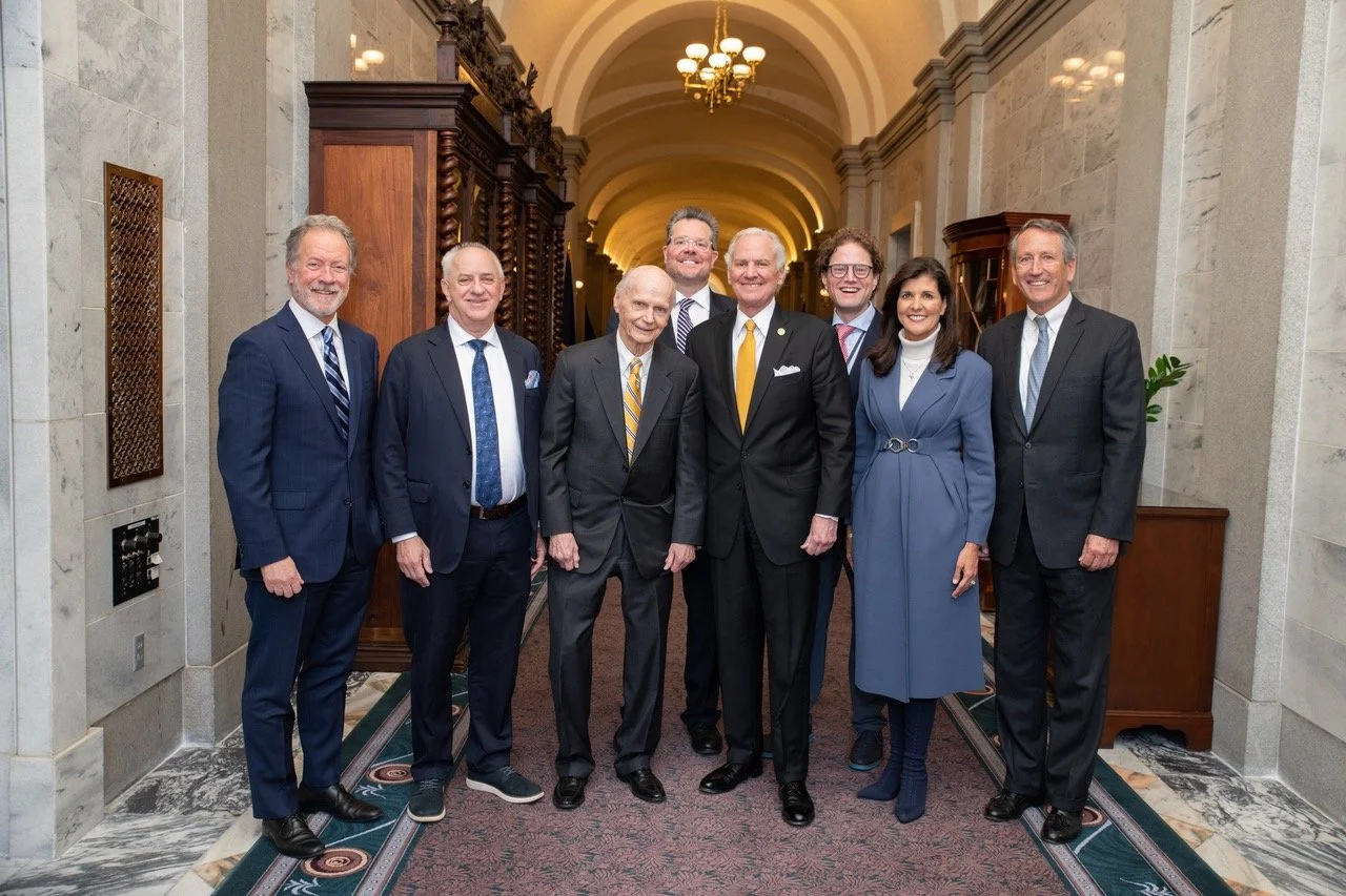 A group of nine diverse business professionals, including men and women, dressed in formal suits, standing together in a hallway with arched ceilings, chandeliers, and historical wooden furniture.