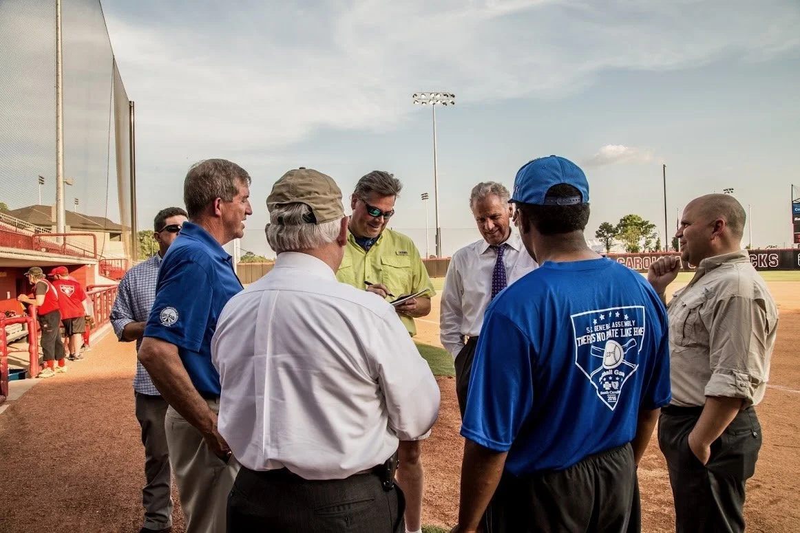 Group of men standing in a circle on a baseball field, some are talking and smiling. The background shows a baseball outfield and stadium lights.