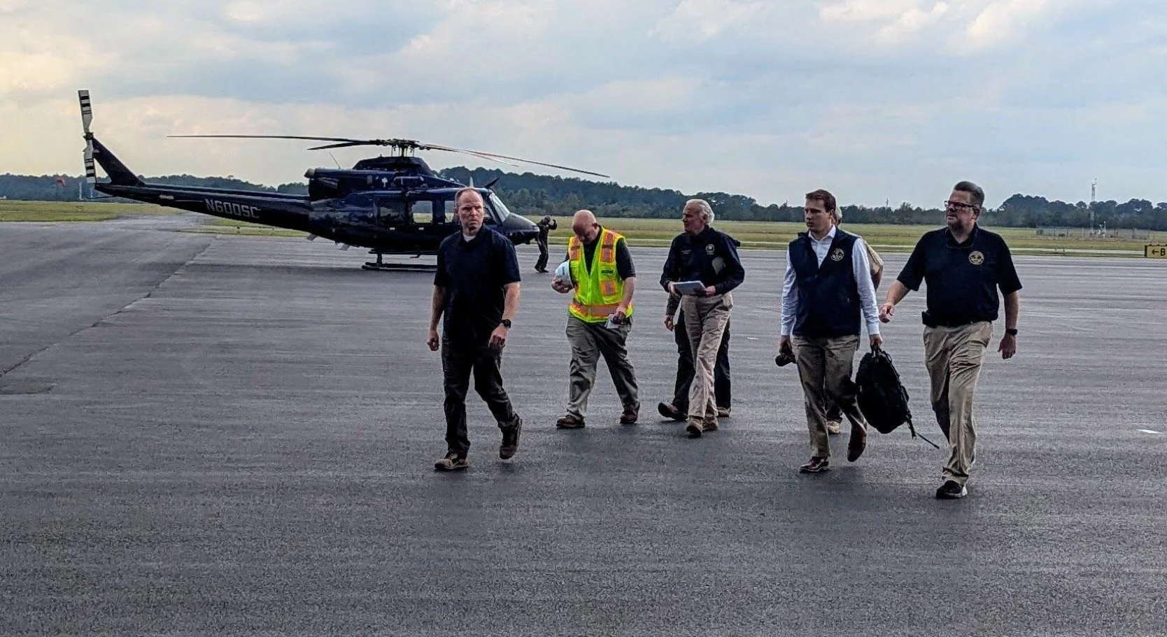 Group of five men walking across an airport tarmac near a black helicopter, with one person carrying a helmet and another with a backpack, under cloudy skies.