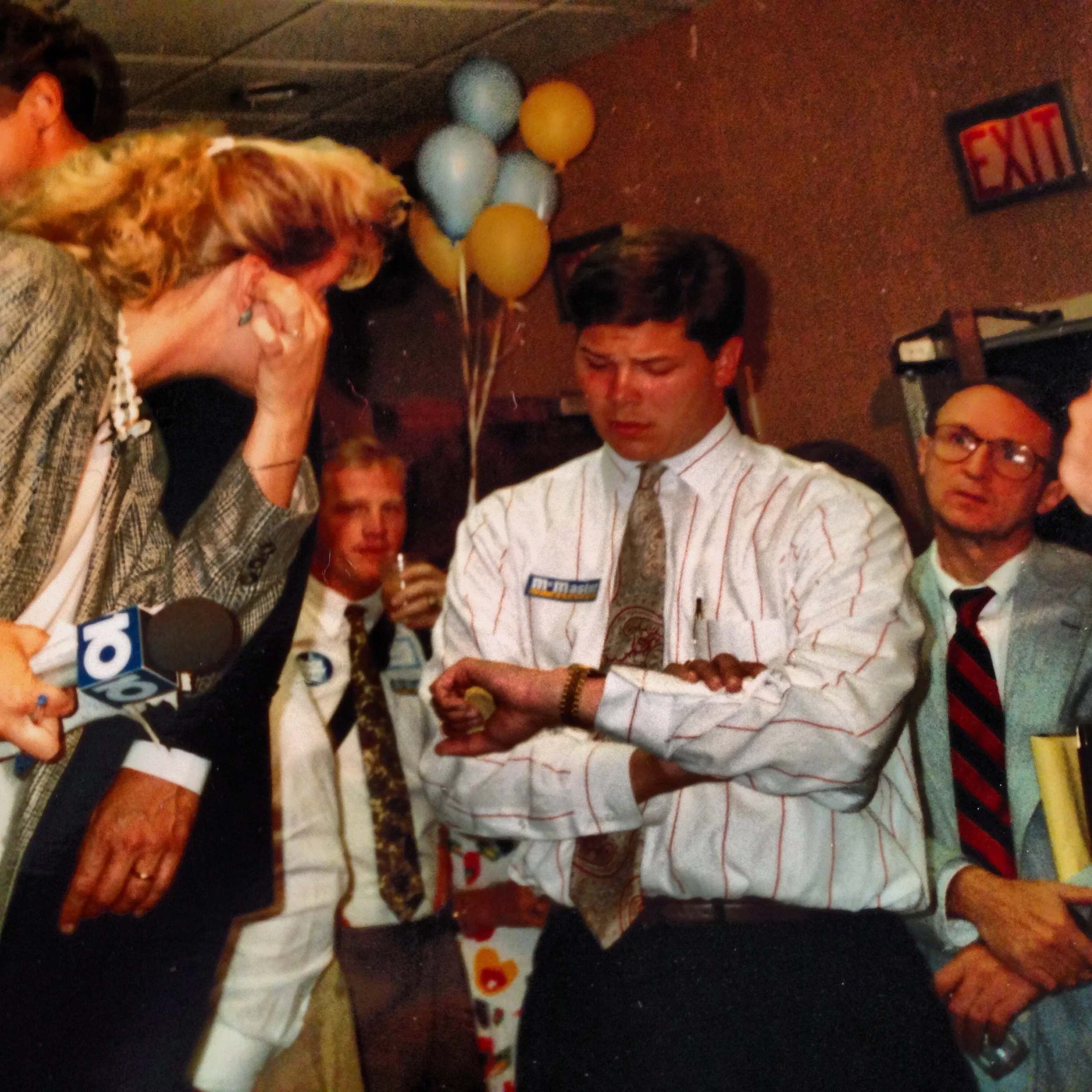 People at a celebration or party, some wearing party hats and balloons in the background, one person appears to be cutting a cake or presenting a gift.