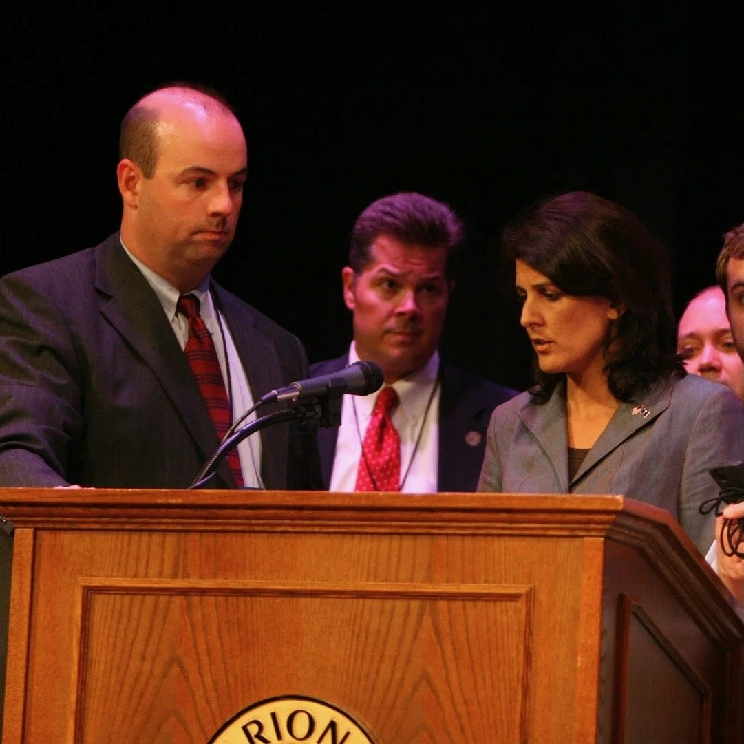 A woman in a gray blazer and a man in a dark suit and red tie are looking down at a wooden podium with a microphone, with two men standing behind them, on a dark background.
