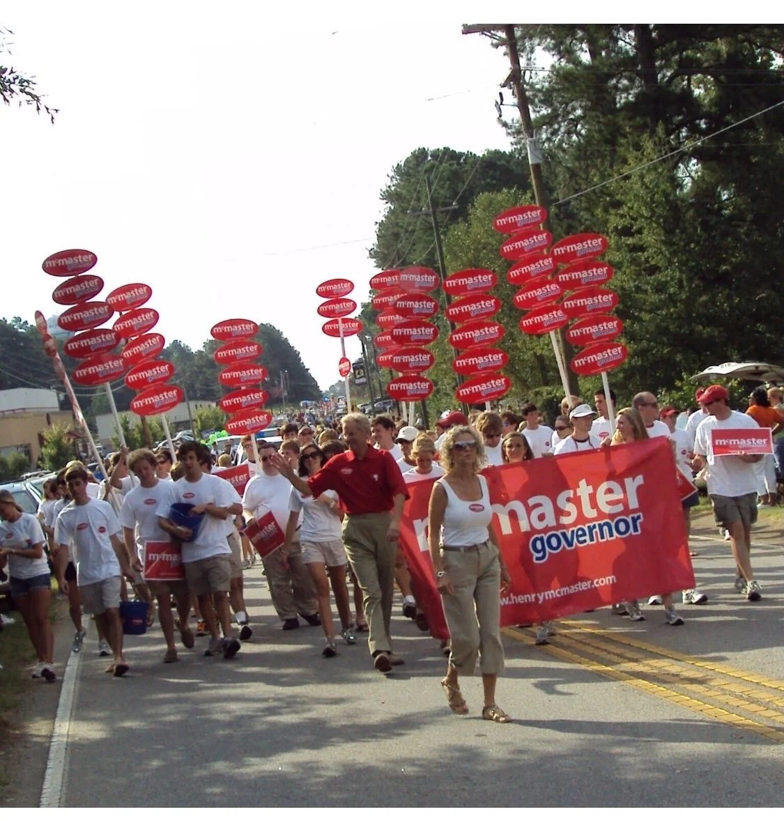 A political rally with supporters marching on a street, holding signs and banners supporting m'master and his campaign for governor.