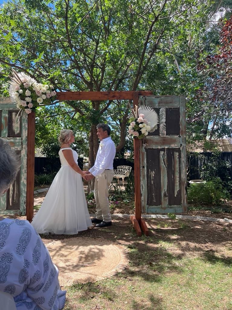 A bride and groom holding hands and standing under a rustic wooden arch decorated with flowers during an outdoor wedding ceremony. Trees and greenery surround them.