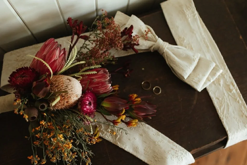 A bouquet of various flowers and a white ribbon, with two wedding rings placed next to it on a dark wooden surface.