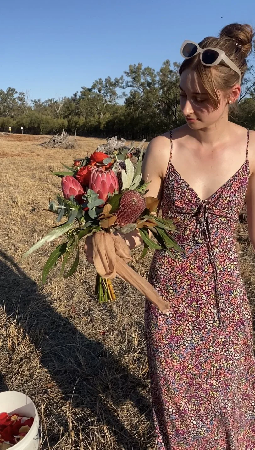 A young woman with sunglasses on her head is holding a bouquet of colorful flowers, including protea, in an open field with trees in the background under a clear blue sky.