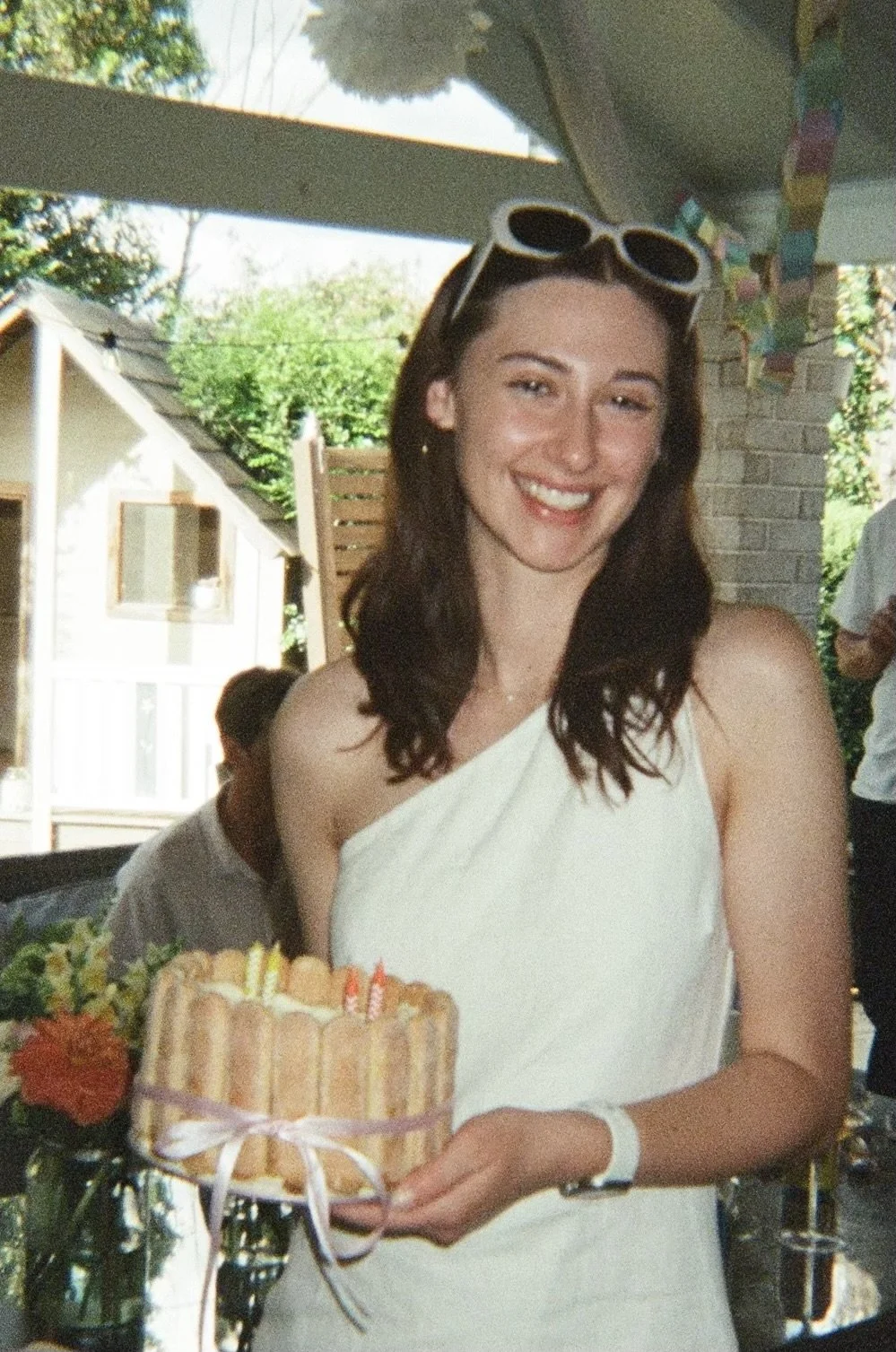 A young woman with brown hair and sunglasses on her head, smiling and holding a birthday cake with candles, wearing a white dress, in an outdoor setting with houses and greenery in the background.