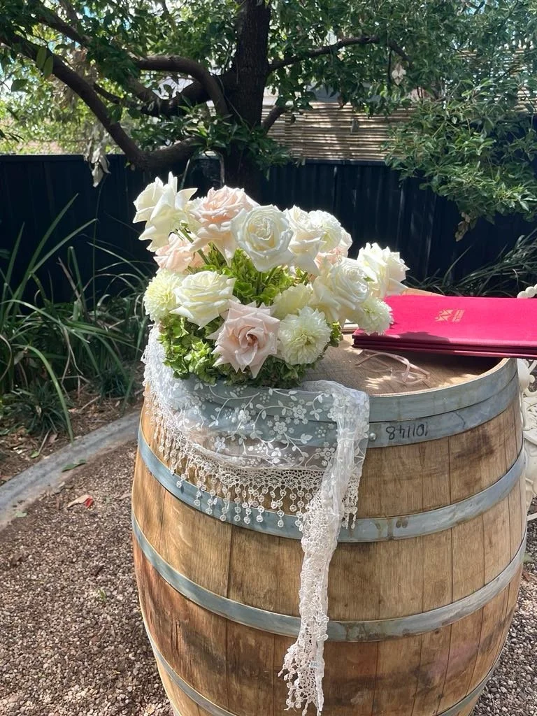 A floral arrangement of white and light pink roses, white dahlias, and other flowers placed on a wooden barrel, with lace fabric draping over the side, outdoors with trees and bushes in the background.