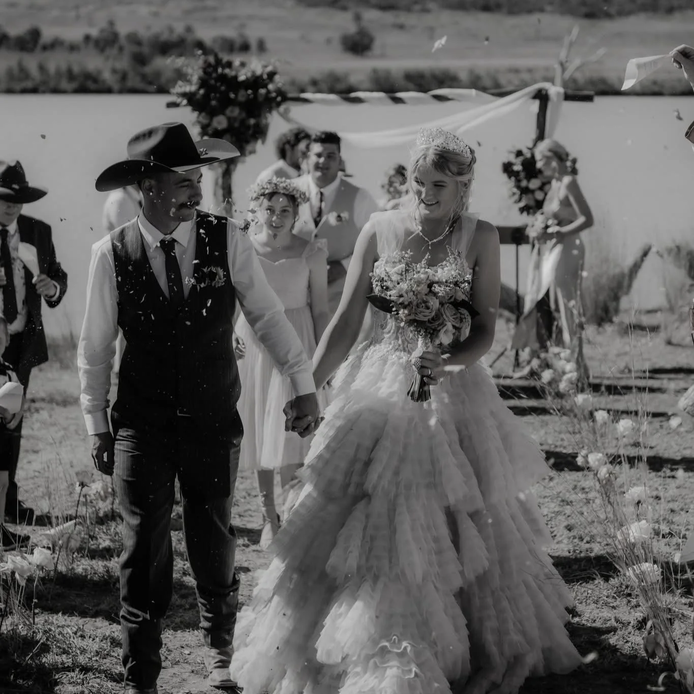 Black and white photo of a wedding scene with a bride and groom holding hands, surrounded by wedding guests outdoors, with decorations and a scenic background.