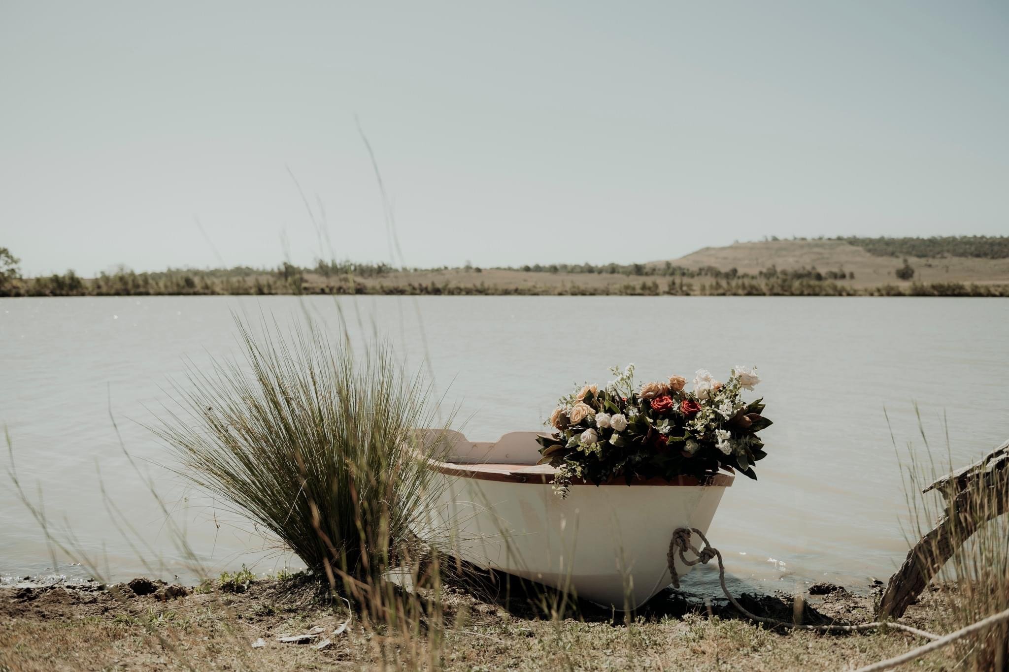 A small boat decorated with a large bouquet of flowers, beside a grassy area on the shore of a body of water, with hills in the background.