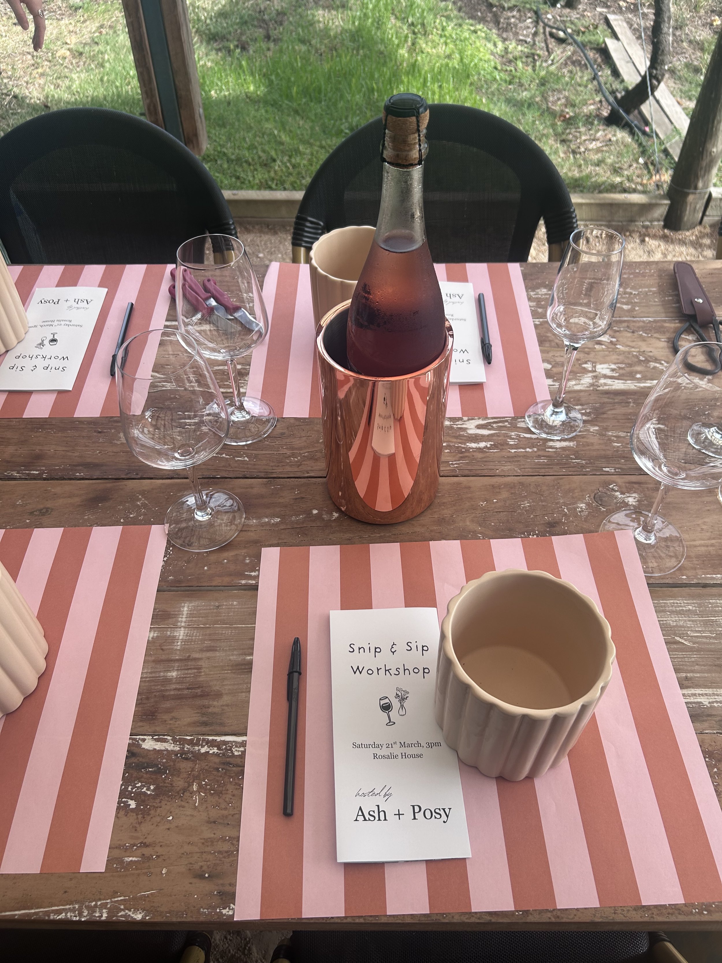 A rustic wooden table set for a wine tasting workshop with pink and red striped placemats, empty wine glasses, a bottle of rosé wine in a copper-colored holder, a cream-colored ramekin, and a printed event program titled 'Snip & Sip Workshop' held on March 21 at Rosalie House, hosted by Ash and Posy.