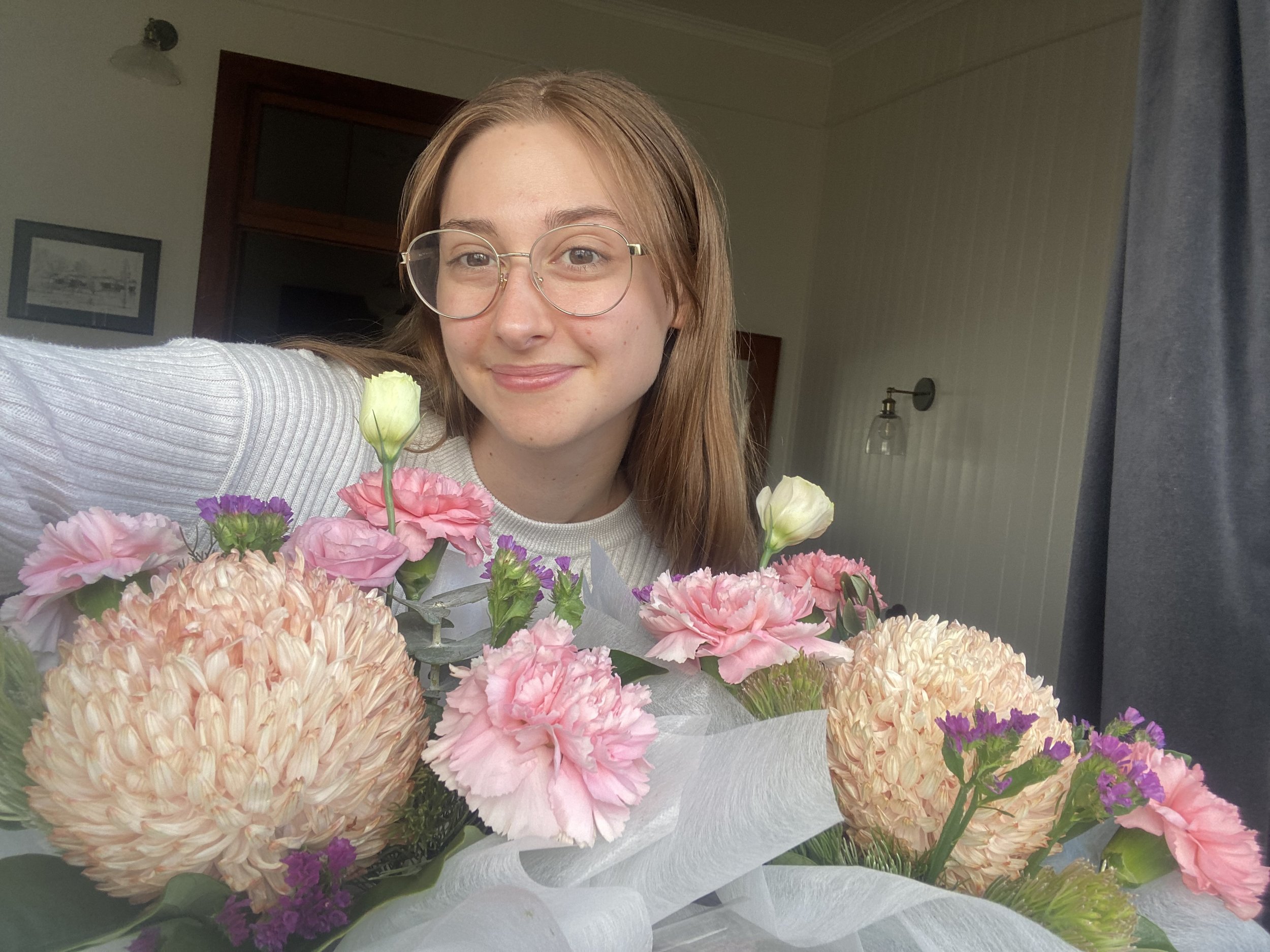 Young woman with glasses smiling and taking a selfie with a large bouquet of pink, white, and purple flowers.