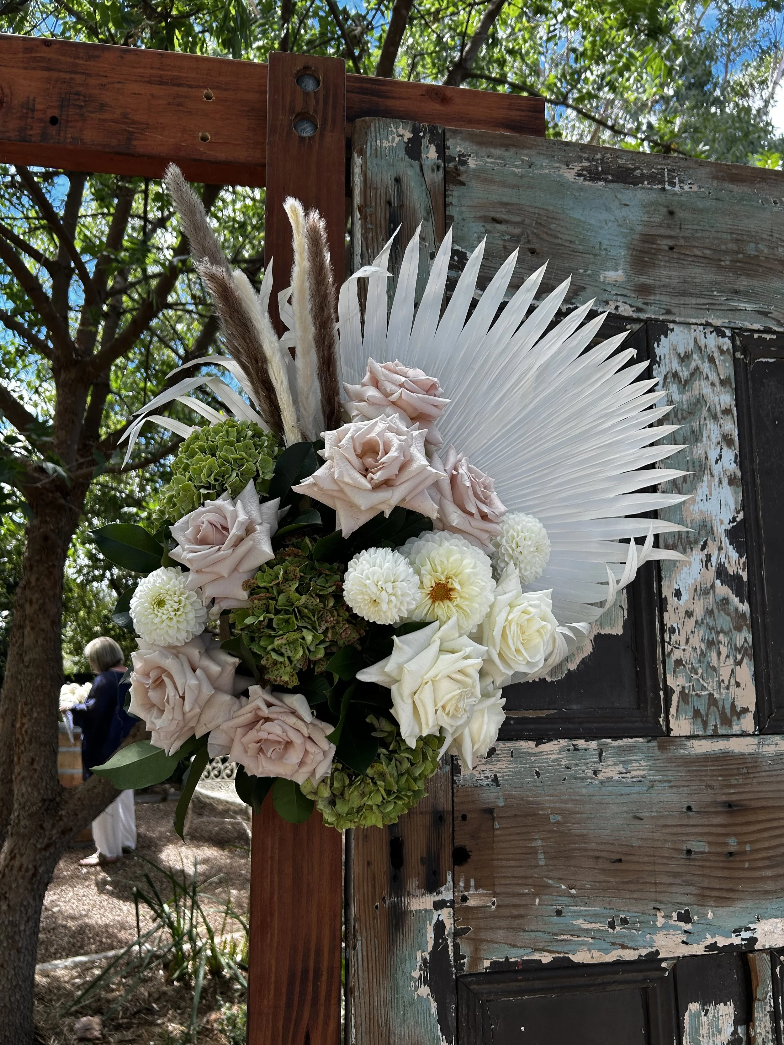 A floral arrangement with pale pink roses, white dahlias, white roses, green hydrangea, and ornamental grasses, attached to a weathered wooden board with a palm leaf backdrop, set outdoors with trees and a person in the background.