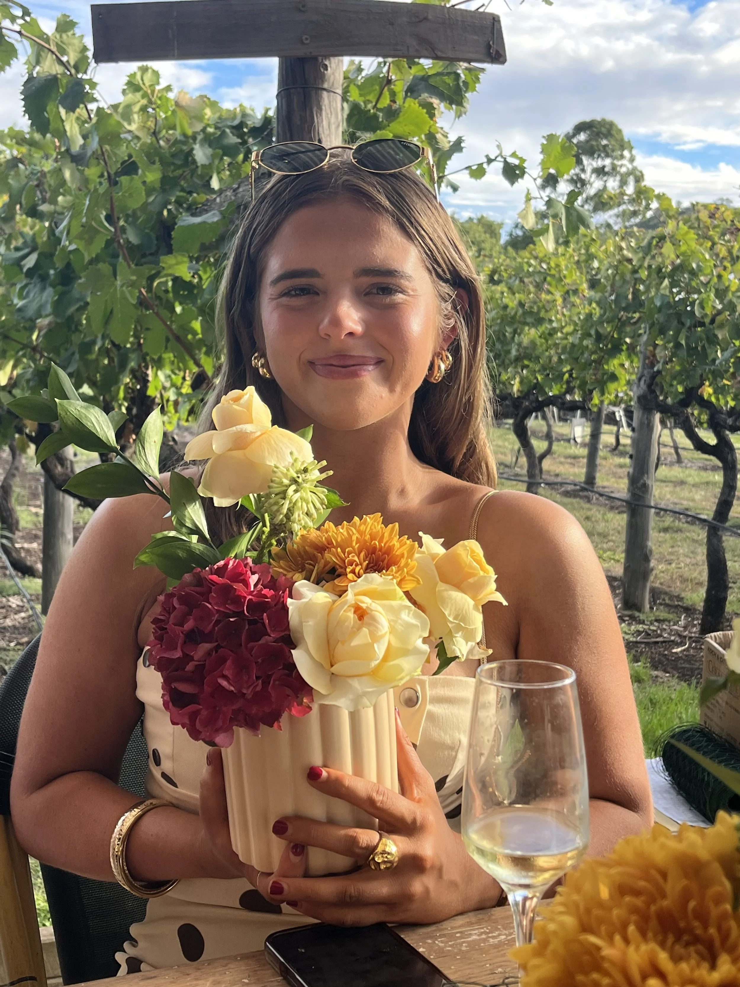 Woman holding a floral bouquet at an outdoor gathering, with wine glass and tablet on table, surrounded by vineyard landscape.